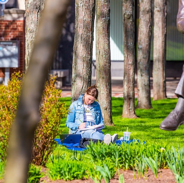 Student leans against a tree and writes in a notebook on Gaebe Common.