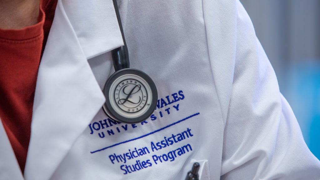 Closeup of a stethoscope and a white coat embroidered with the words, “Johnson &amp; Wales University Physician Assistant Studies Program.”