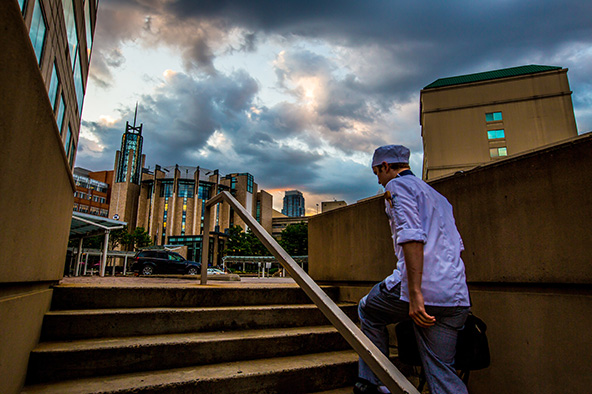 A student walking up steps to JWU Charlotte, with a sunset over the main campus building. The sky is dark and full of beautiful colors.