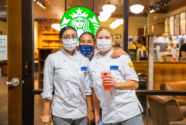 three students standing in front of the on-campus starbucks, posing for the camera and wearing face masks.