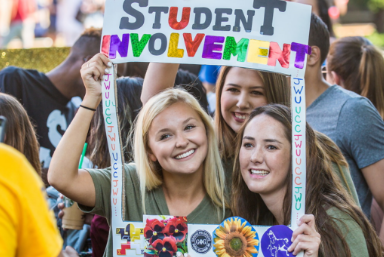 3 female students posing for a picture. Holding a frame that reads "Student Involvement"