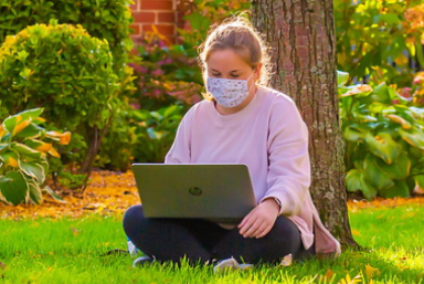 female student sitting outside by a tree on her laptop wearing a mask