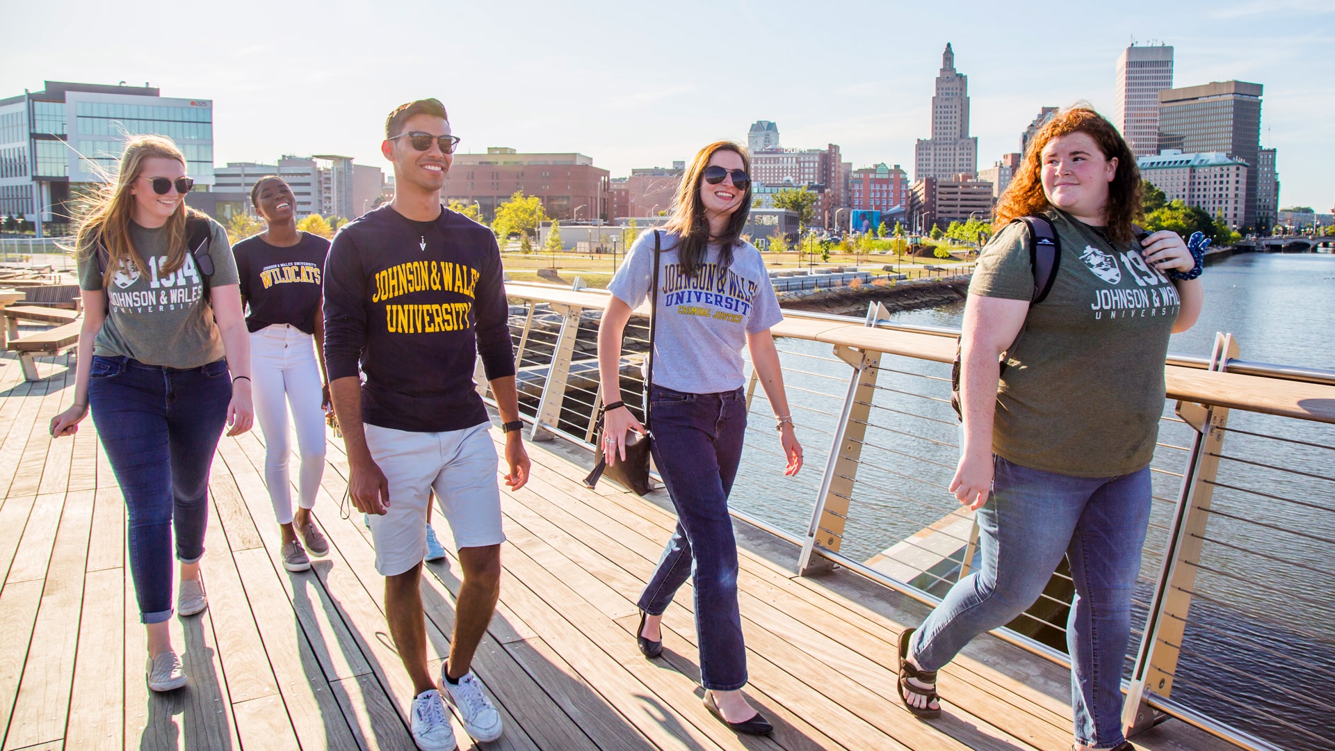 Group of JWU Providence students crossing the Downcity pedestrian bridge with the city skyline in the background.