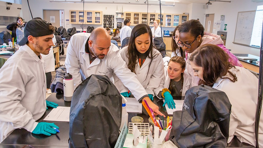 an engaged biology class of students and instructor wearing white coats interact in a lab together
