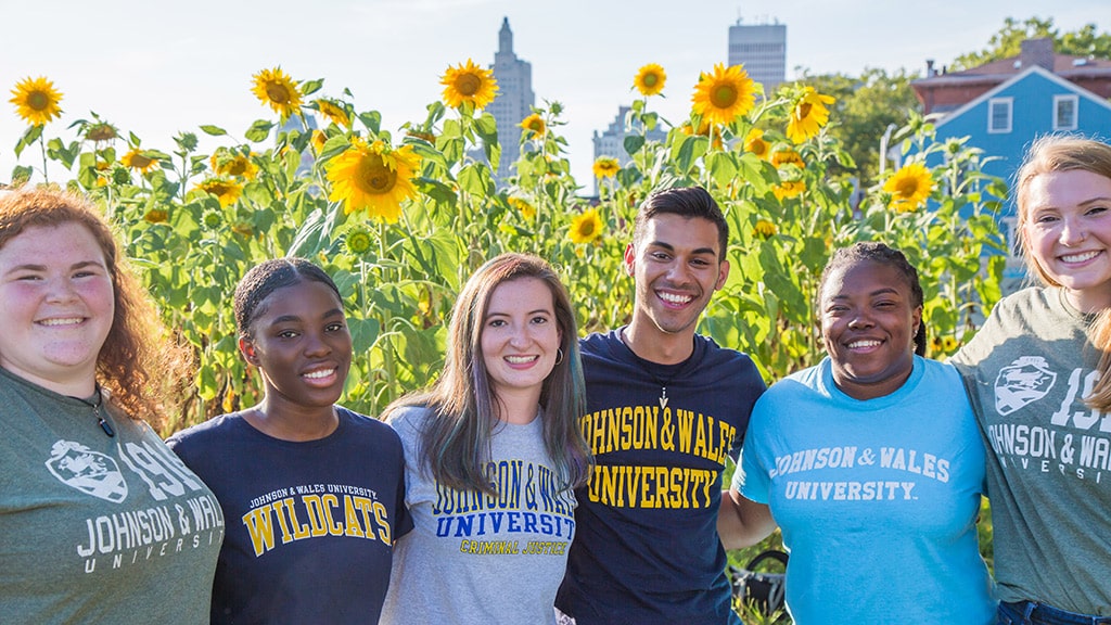 a group of JWU Providence students smile for a photo by a field of sunflowers in front of the city skyline