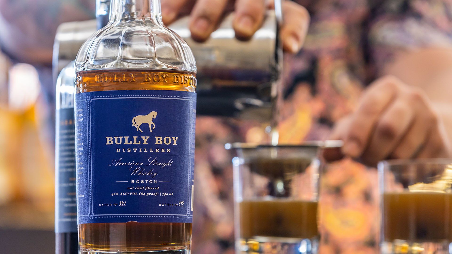 A bottle of Bully Boy whiskey in the foreground with a bartender pouring drinks through a strainer in the background