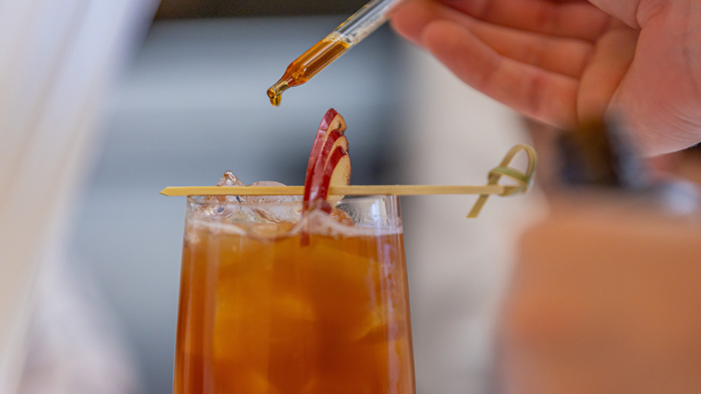 A dropper places drops of bitters into an amber colored drink topped with apple slices