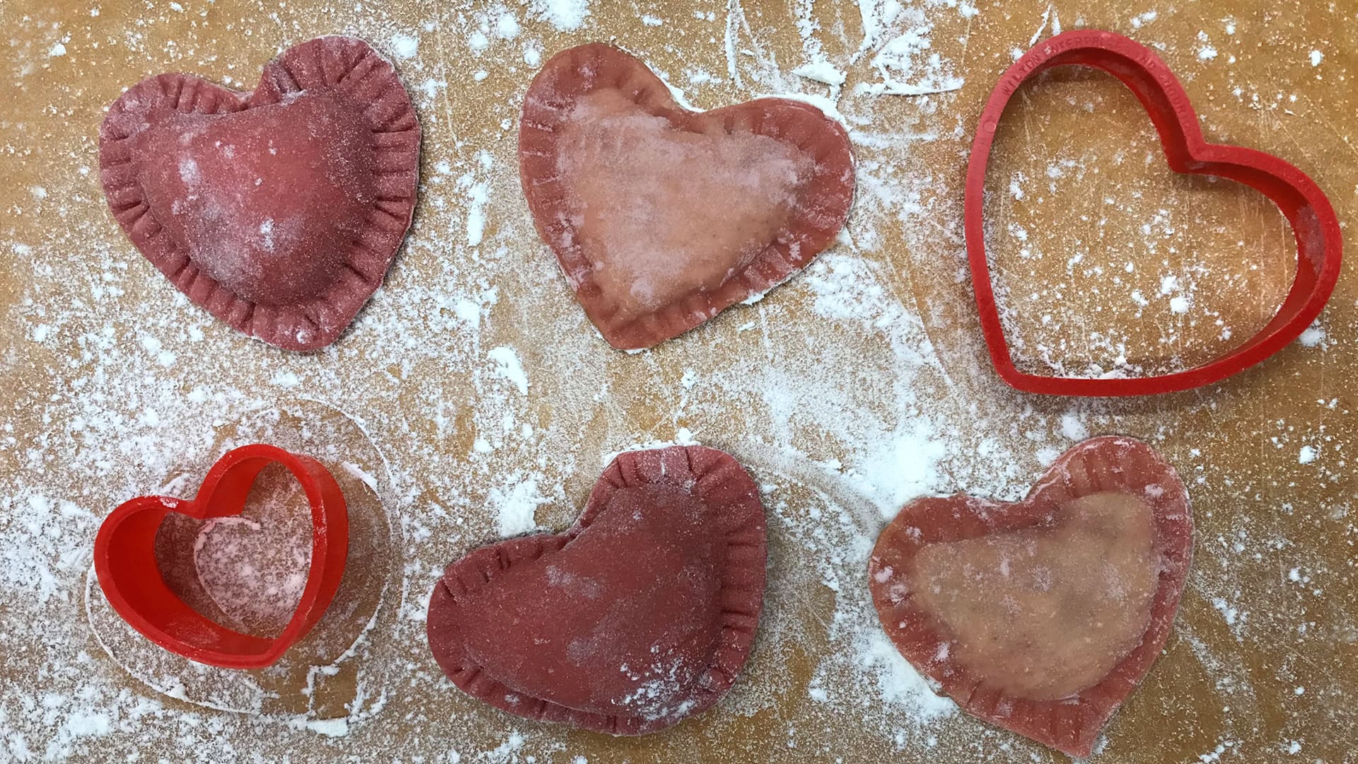 Heart-shaped ravioli pasta and heart-shaped cookie cutters arranged in a row on a floured cutting board.