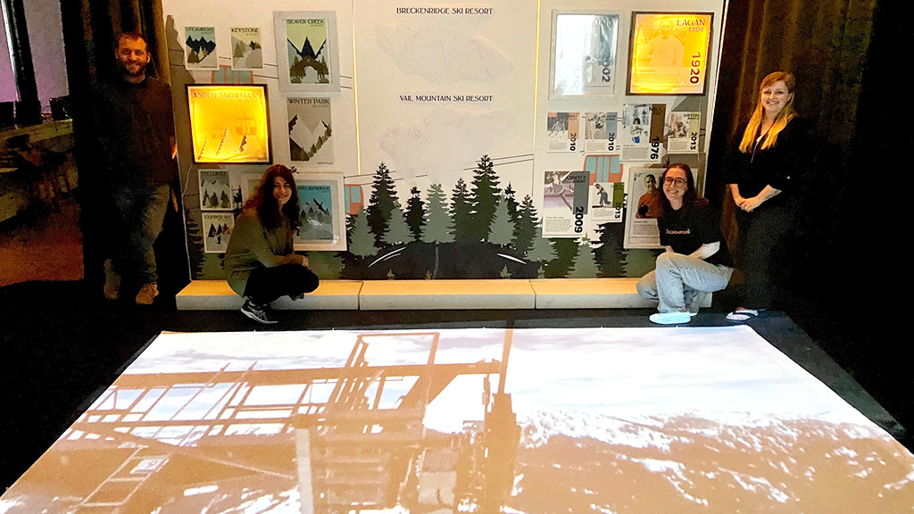 Students pose in front of their project about a ski resort with a projection on the floor in front of them