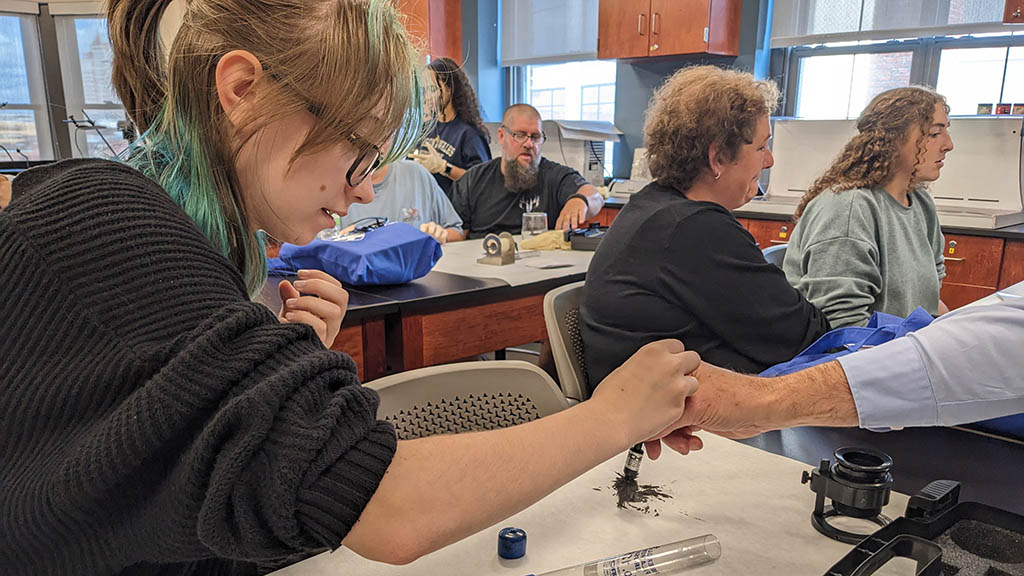 a photo of a student attending Experience Day and having her hand guided in using magnetic powder to reveal a fingerprint