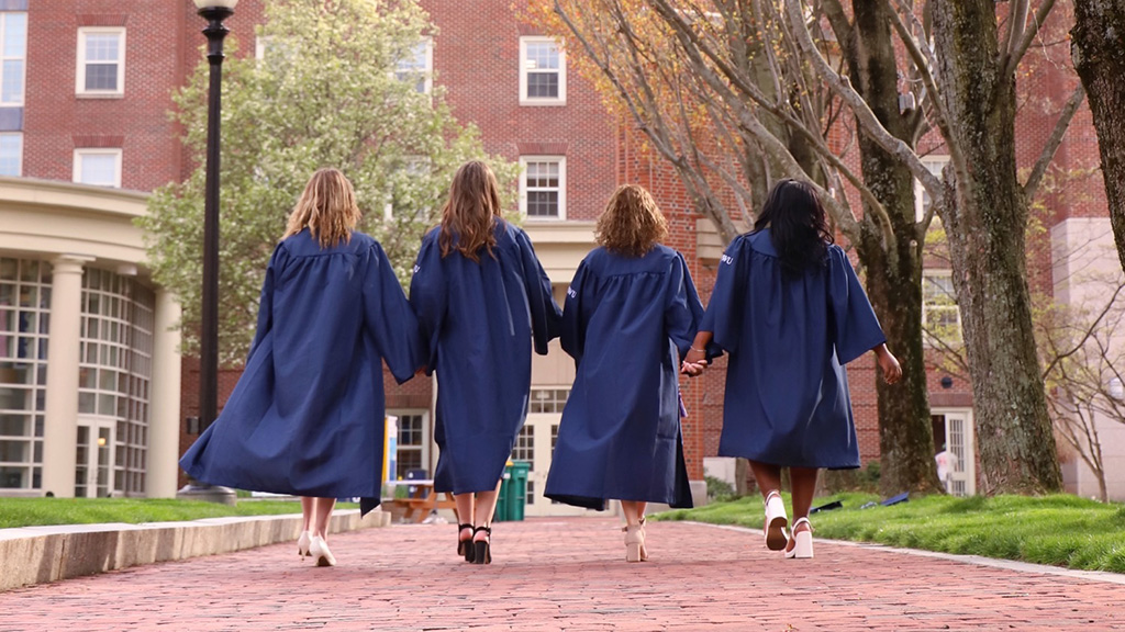 Julia and three friends walking through Gaebe Commons with their graduation gowns on.