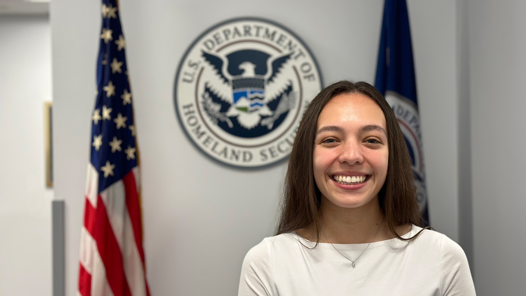 a photo of Sofia Tamayo '24 smiling while posing in front of a Department of Homeland Security seal and flags
