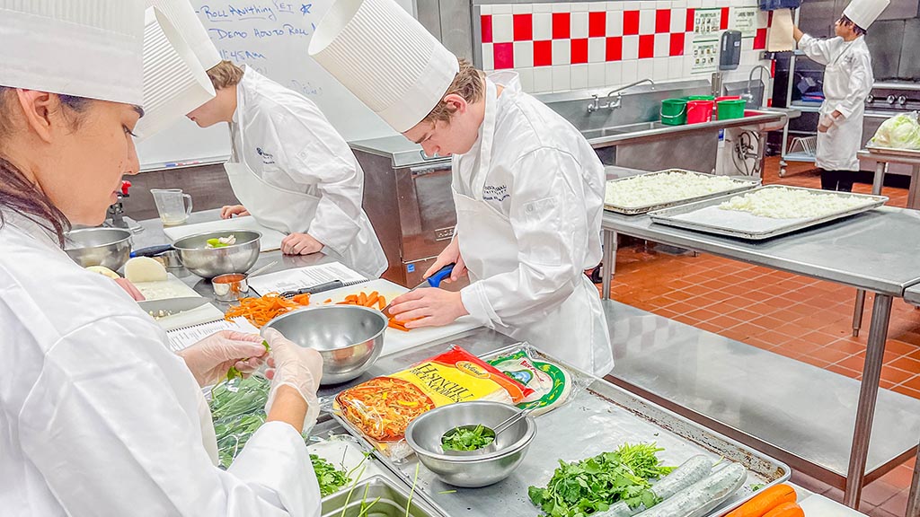 Students practice knife skills by chopping carrots at a prep station.