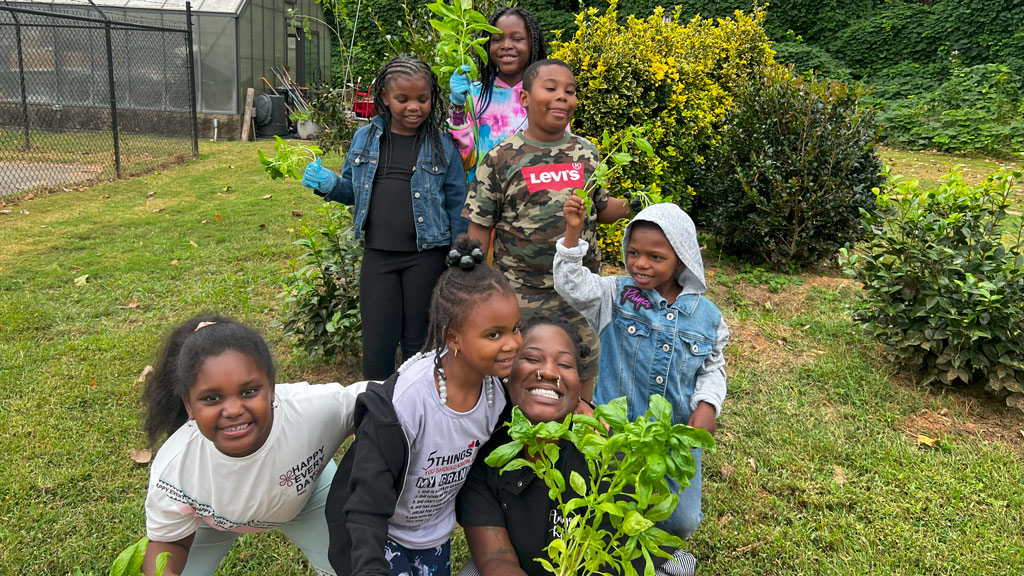 a woman in the forefront grins and clutches herbs while surrounded by a group of children outdoors