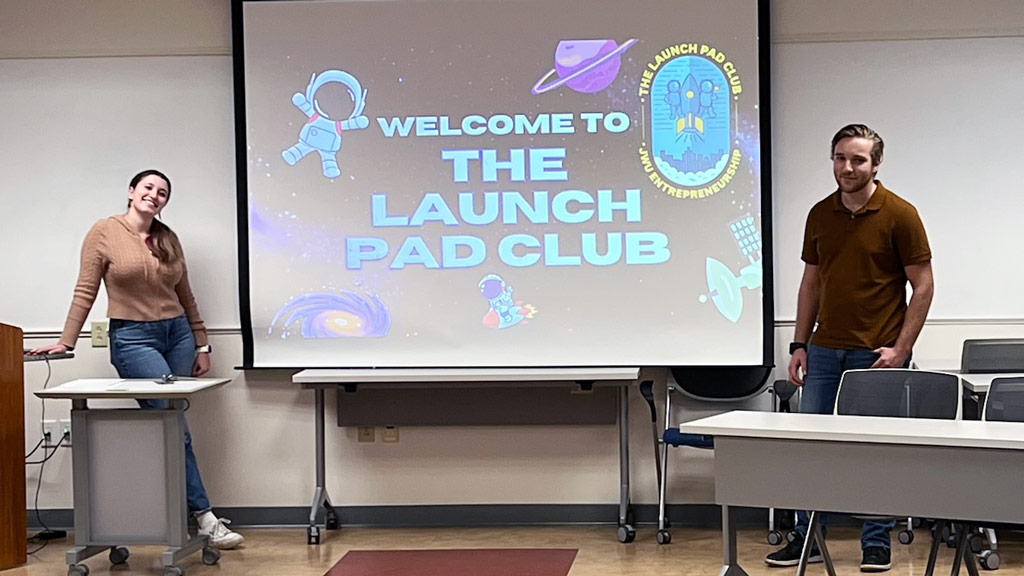 two students pose on either side of a screen titled "Welcome to The Launch Pad Club"