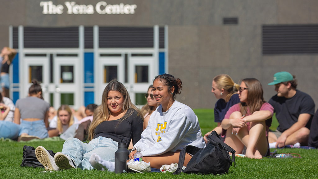 Two students sit on grass with other students in the background