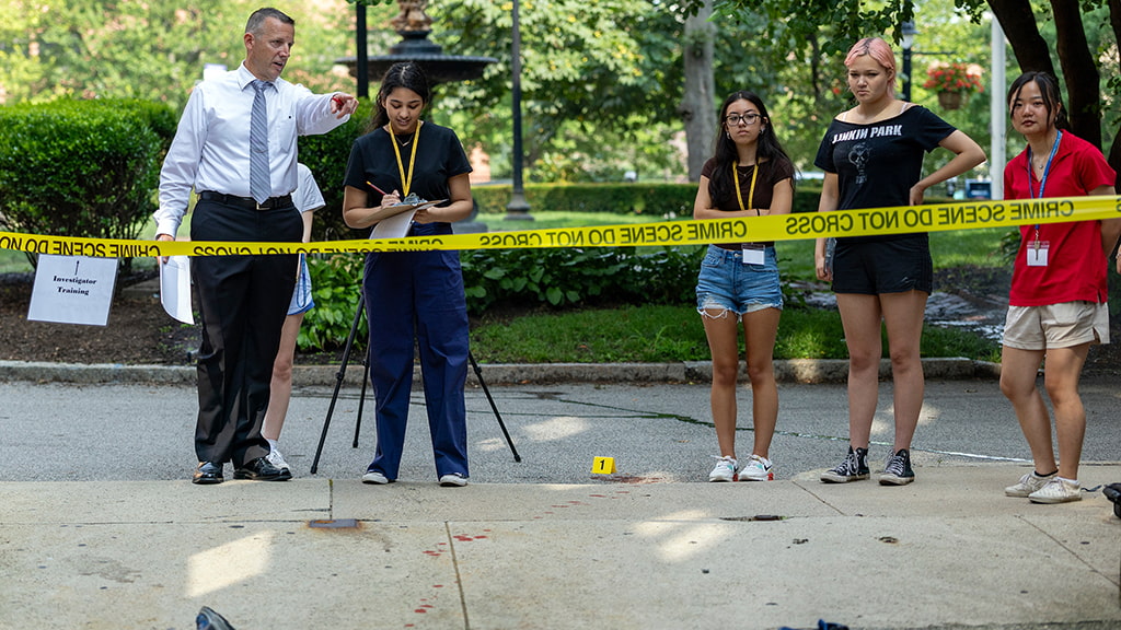 At a staged crime scene, a professor points past caution tape as students watch and take notes