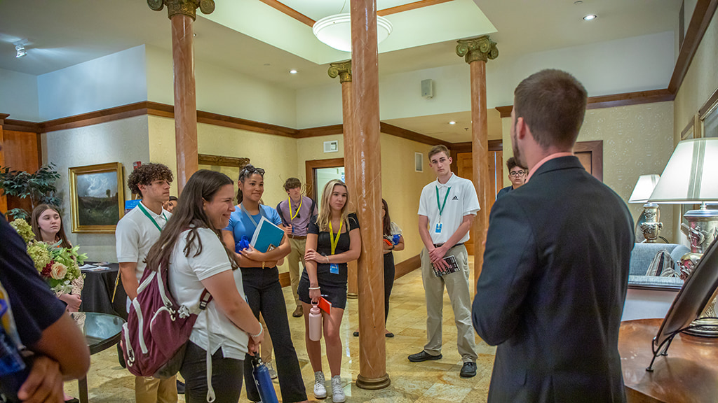 A group of students watch the manager of a hotel lecture in the lobby