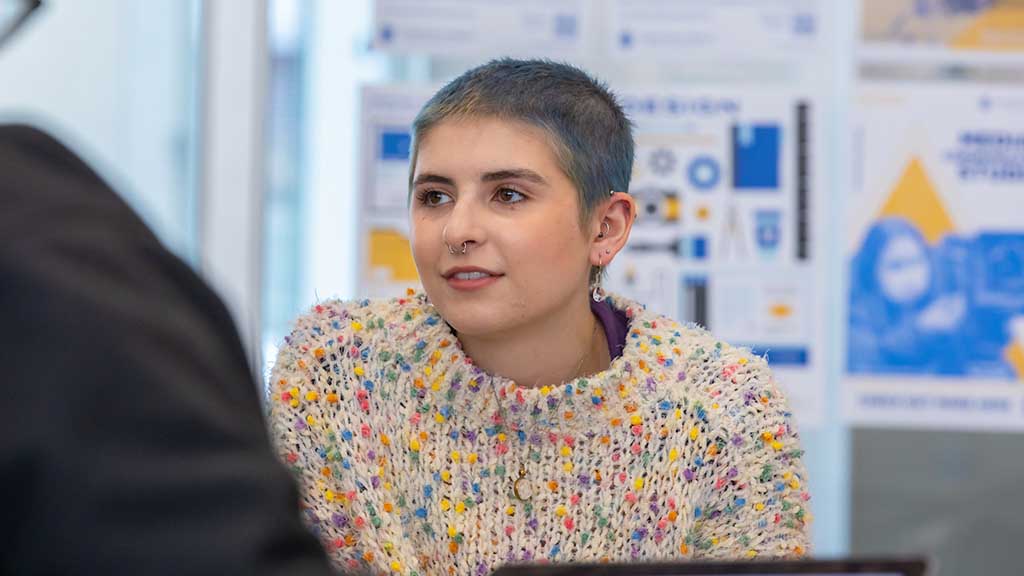 Person with a short haircut wearing a colorful knitted sweater, listening attentively in a meeting with graphic designs in the background.