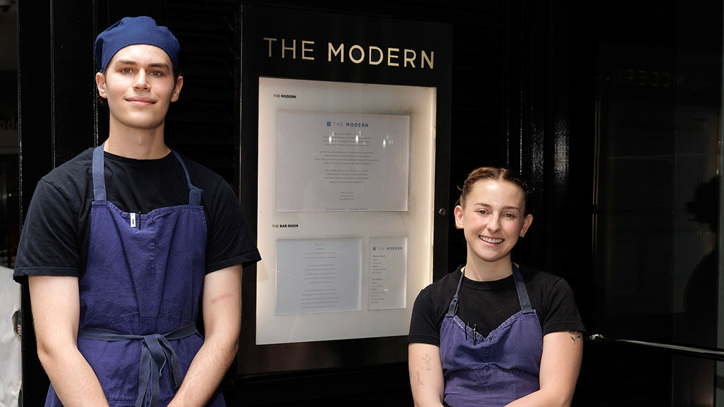 Daron Sklar '27 (left) and Charlotte Brecher ’18 (right), both wearing black shirts and denim aprons as the pose by the front door of The Modern in NYC.