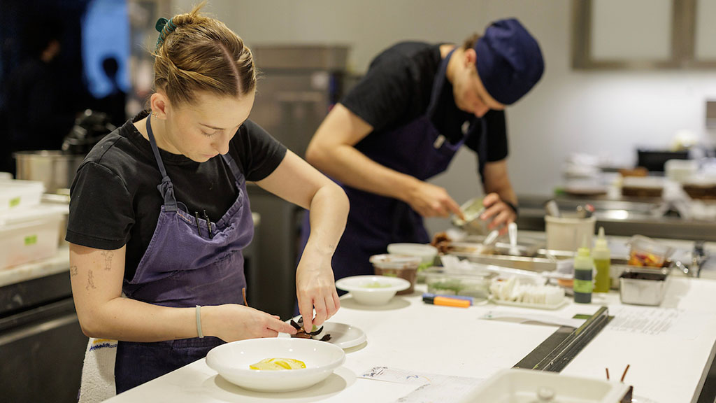 Charlotte Brecher ’18, executive pastry chef at The Modern (left); Daron Sklar '27, pastry line cook at The Modern (right); both wearing black shirts and denim aprons as they plate desserts.