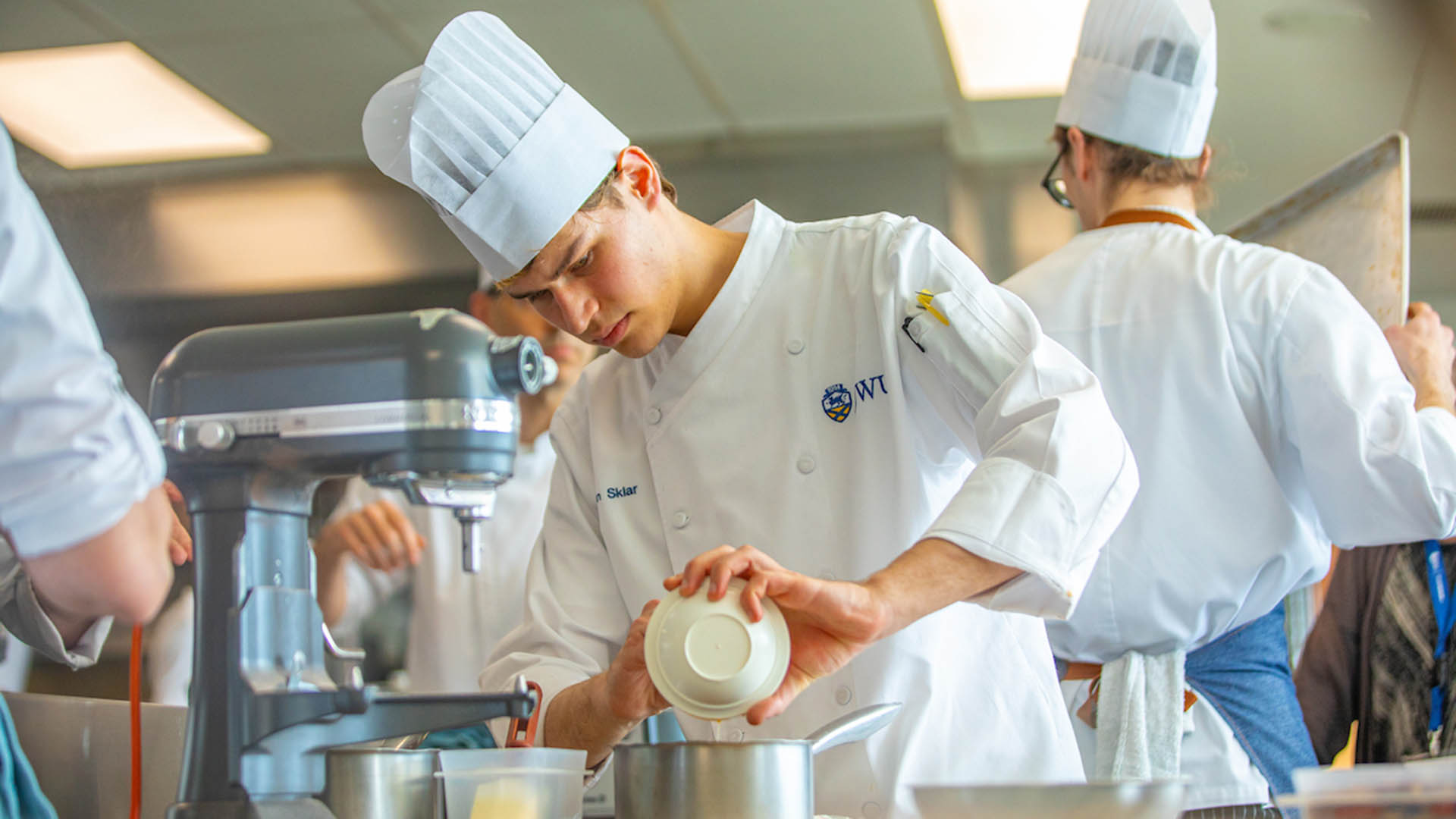 Daron Sklar '27 working at a stand mixer in a white chef hat and coat.